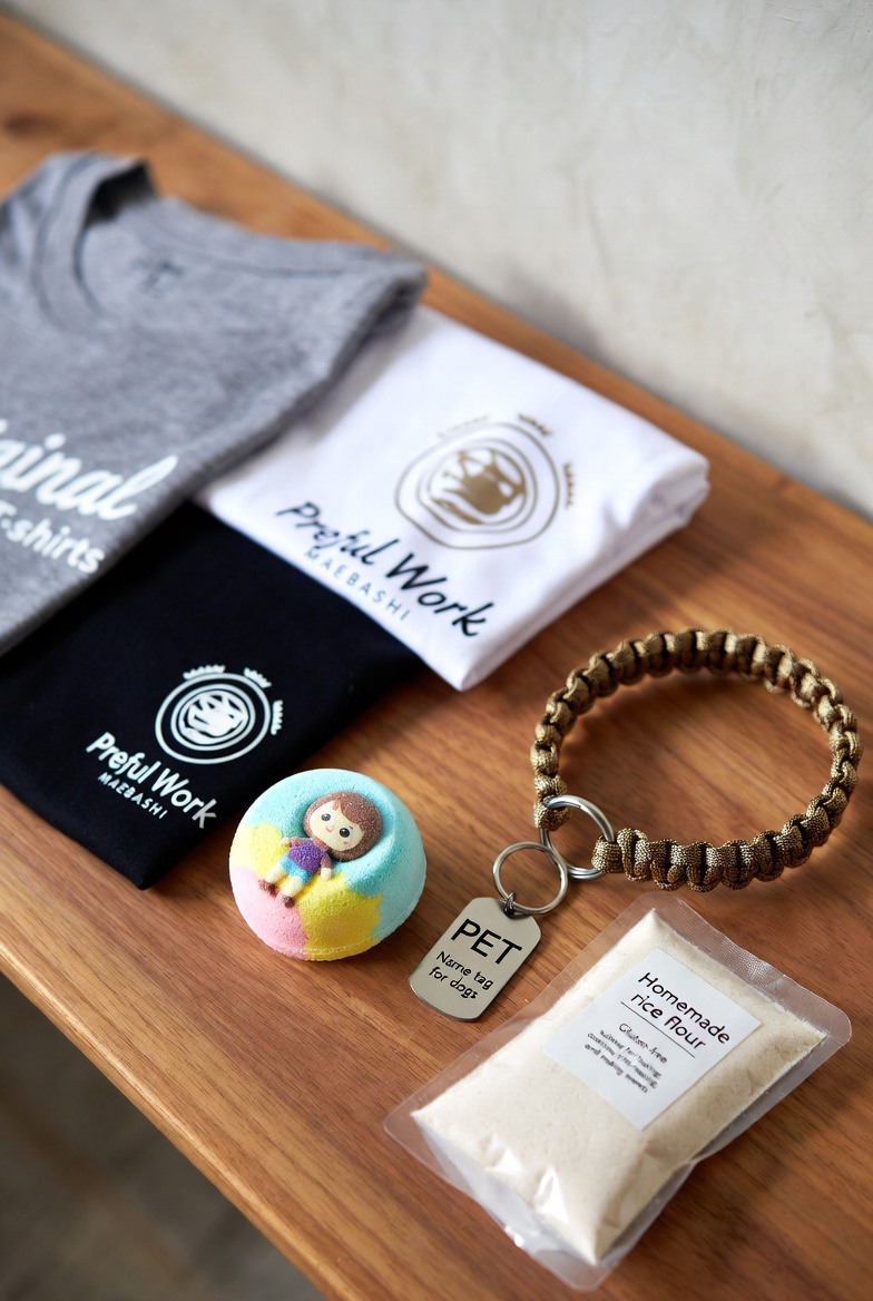 プレフルワーク前橋 商品一覧 — Tシャツ・バスボム・ドッグタグ・パラコードマクラメ首輪・自家製米粉（木製テーブルに並べた商品写真）